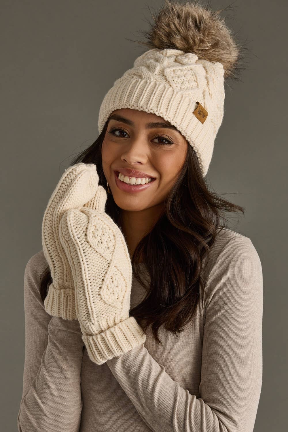 Woman wearing a cream knit beanie and mittens against a gray background
