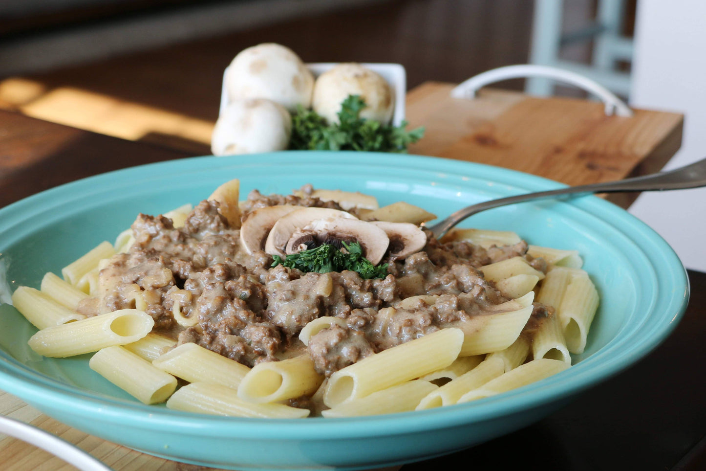 Pasta dish with ground meat, mushrooms, and herbs in a teal bowl on a wooden table.