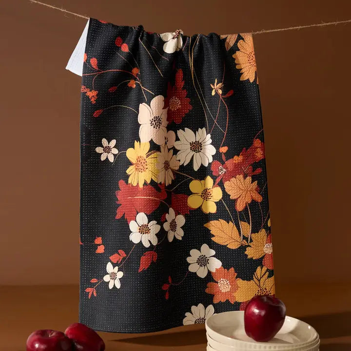 A black kitchen towel with a colorful floral and leaf pattern hangs from a string on a clothesline. The pattern features white, yellow, orange, and red flowers and leaves.