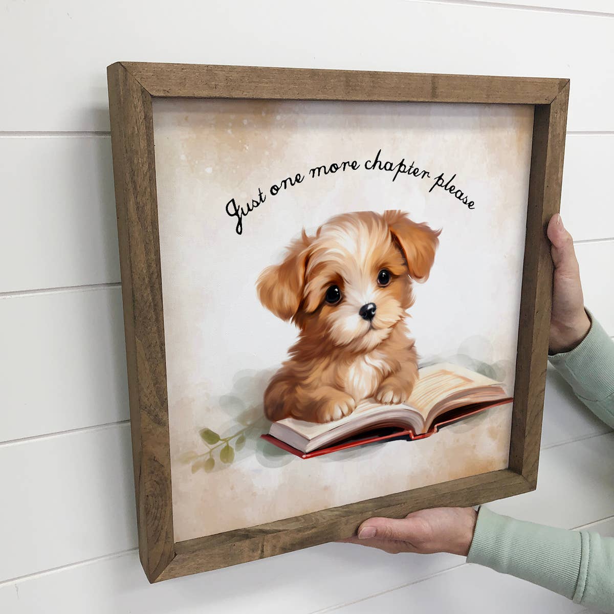 Framed artwork of a puppy reading a book with text 'Just one more chapter please' held by a person.
