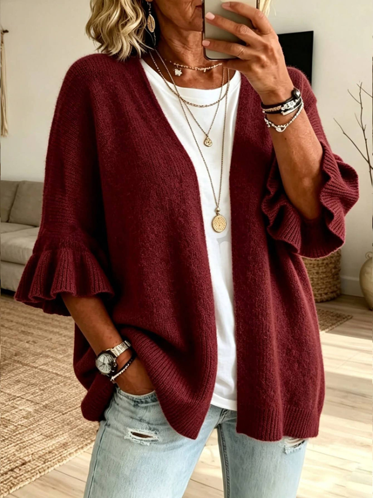 A woman wearing a burgundy open-front knit cardigan with ruffled three-quarter sleeves, layered over a white t-shirt and light-wash distressed jeans, taking a mirror selfie in a living room setting.