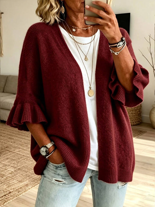 A woman wearing a burgundy open-front knit cardigan with ruffled three-quarter sleeves, layered over a white t-shirt and light-wash distressed jeans, taking a mirror selfie in a living room setting.