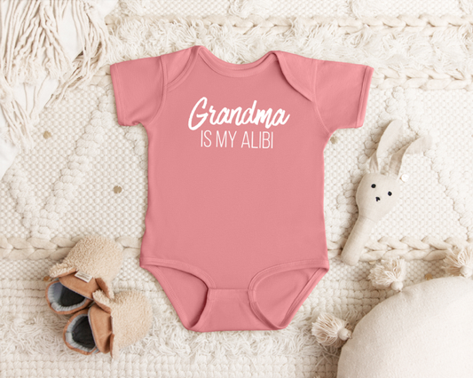 Pink baby onesie with 'Grandma is my alibi' text on a textured surface with toys.