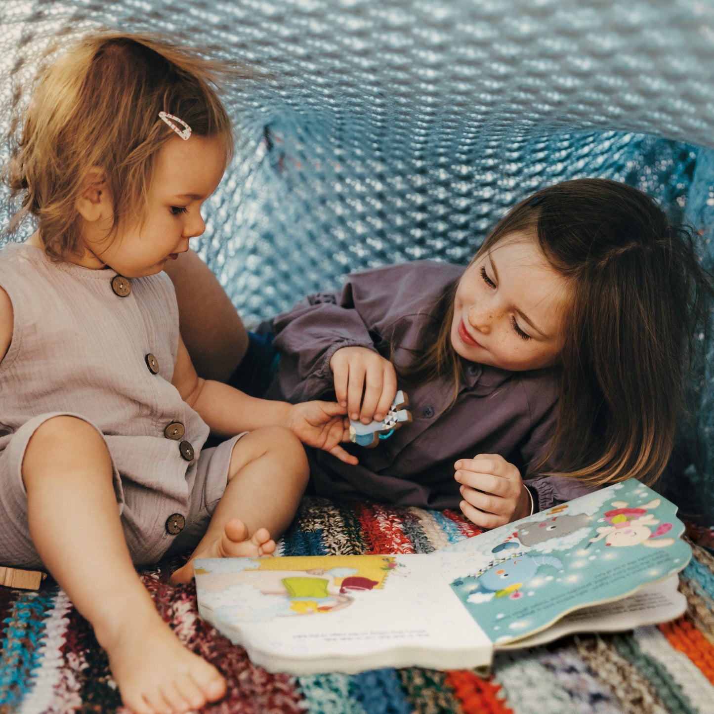 Two young girls sitting on a colorful blanket, looking at a book together.