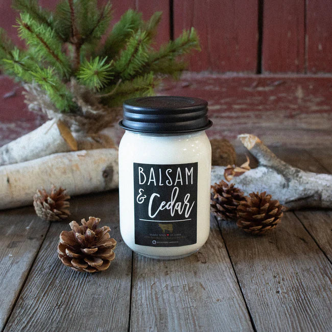 Candle labeled 'Balsam & Cedar' on a wooden surface with pinecones and a small tree branch.