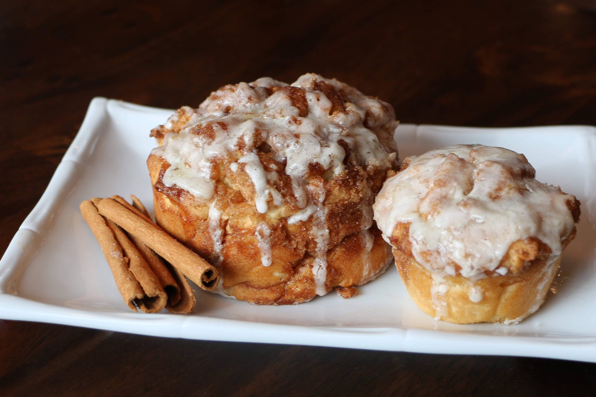 Two cinnamon rolls with icing on a white plate with cinnamon sticks on a wooden surface