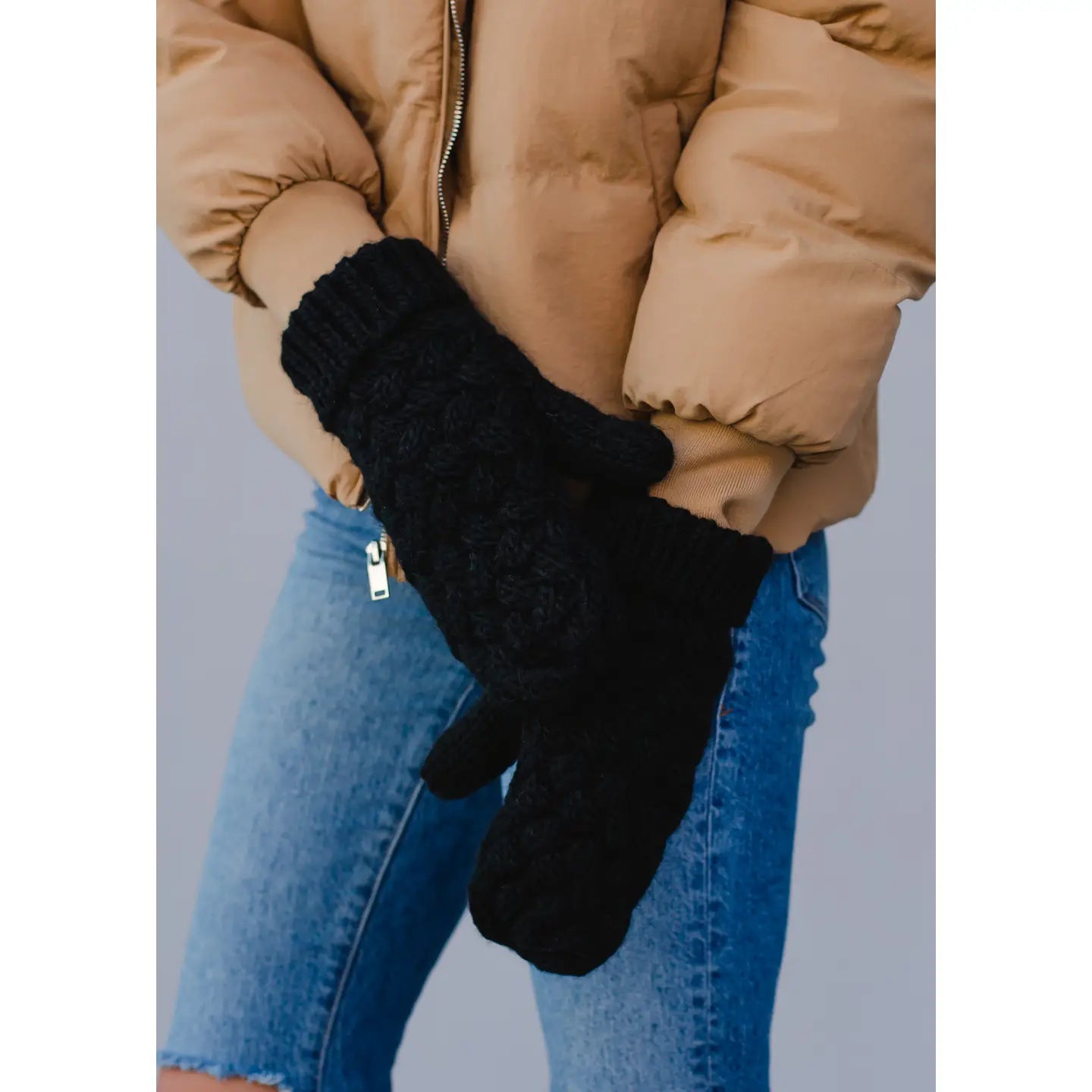 Person wearing black knit gloves with a beige jacket and blue jeans on a light gray background