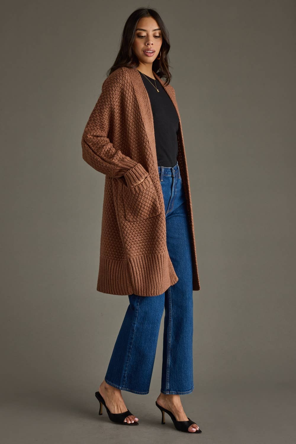 Woman wearing a brown cardigan, black top, and blue jeans against a gray background