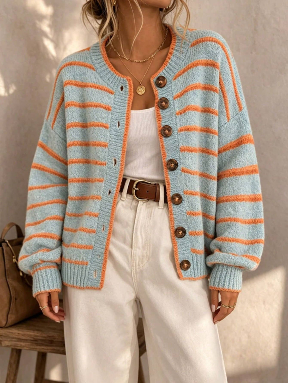 A woman wearing a light blue and orange striped knit cardigan with brown buttons, styled with a white tank top, white pants, and a brown belt.