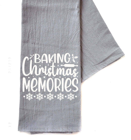 A gray kitchen tea towel with white printed text reading 'BAKING Christmas MEMORIES' and snowflake designs, laid flat on a white background.