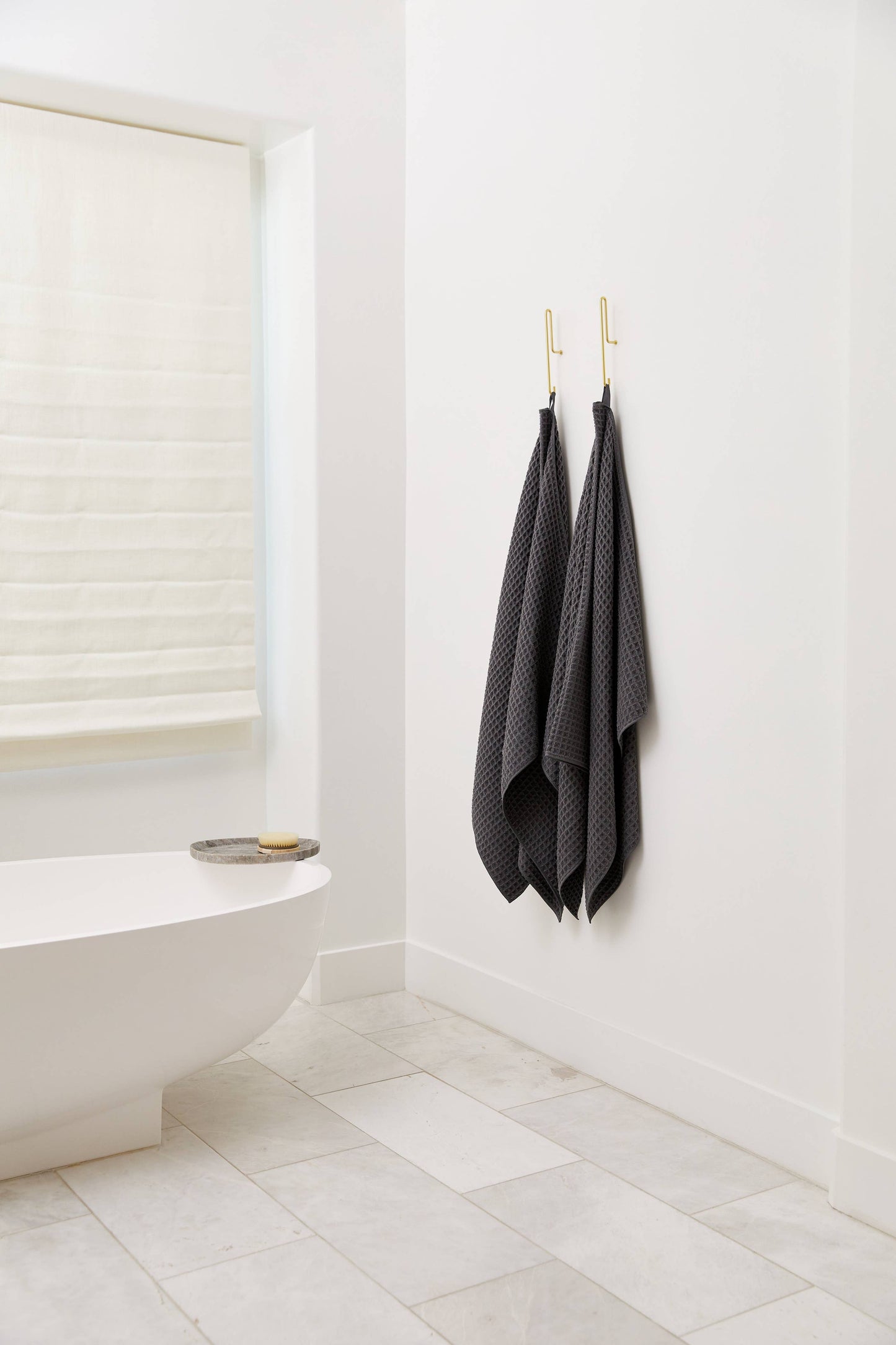 Geometry Waffle Bath Towel Charcoal Two dark gray towels hanging on a hook in a bathroom with a white bathtub and tiled floor.