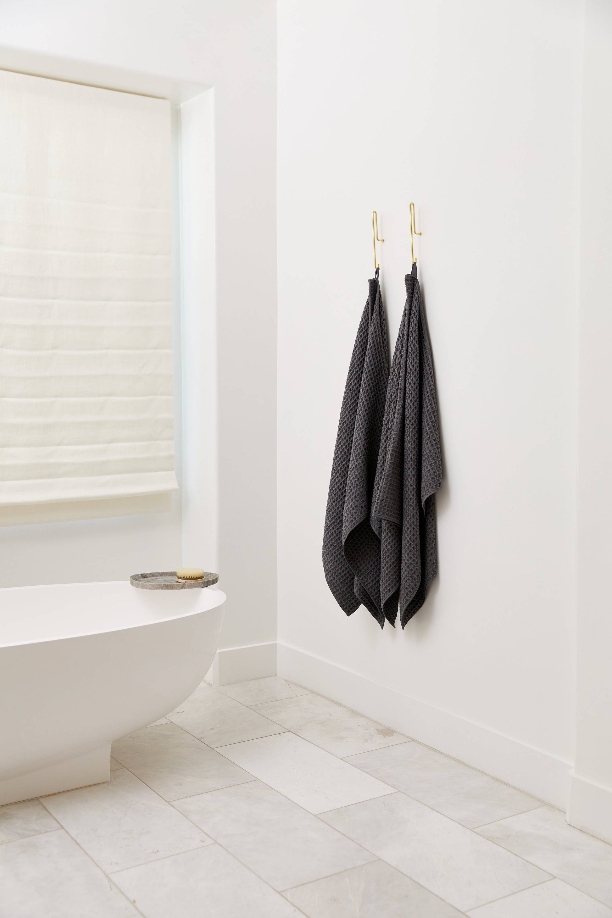 Geometry Waffle Bath Towel Charcoal Two dark gray towels hanging on a hook in a bathroom with a white bathtub and tiled floor.