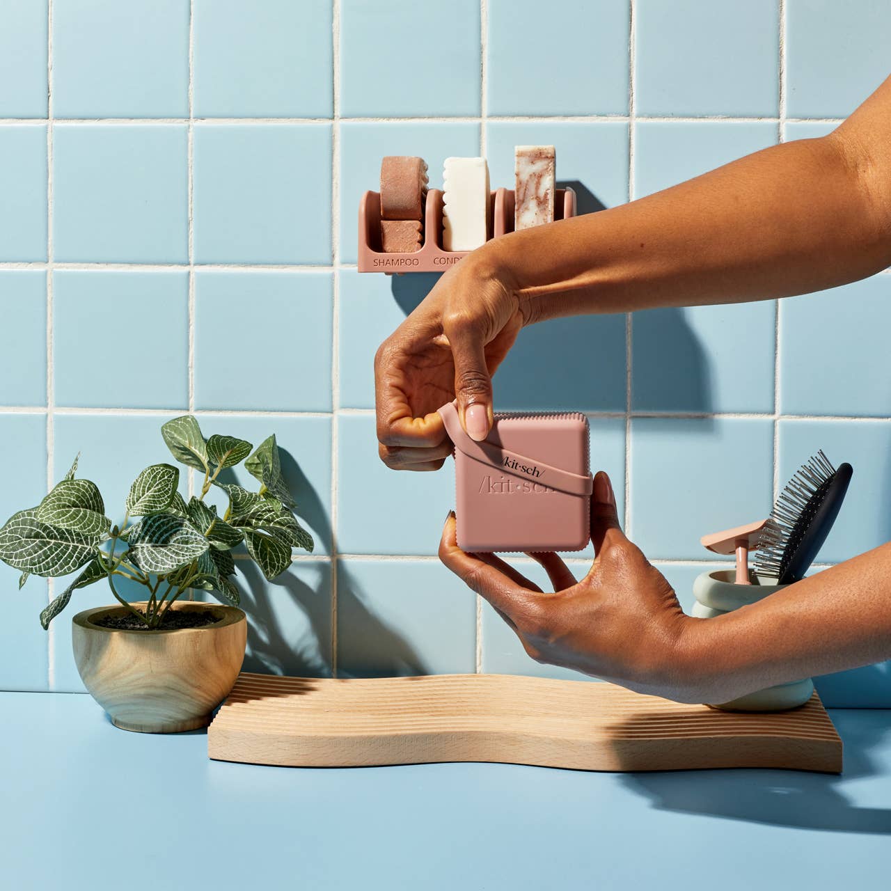 Person holding a pink travel case with a plant and grooming tools on a blue tiled wall background