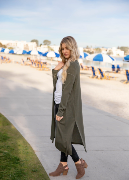Woman wearing an olive green cardigan on a beach with blue umbrellas in the background