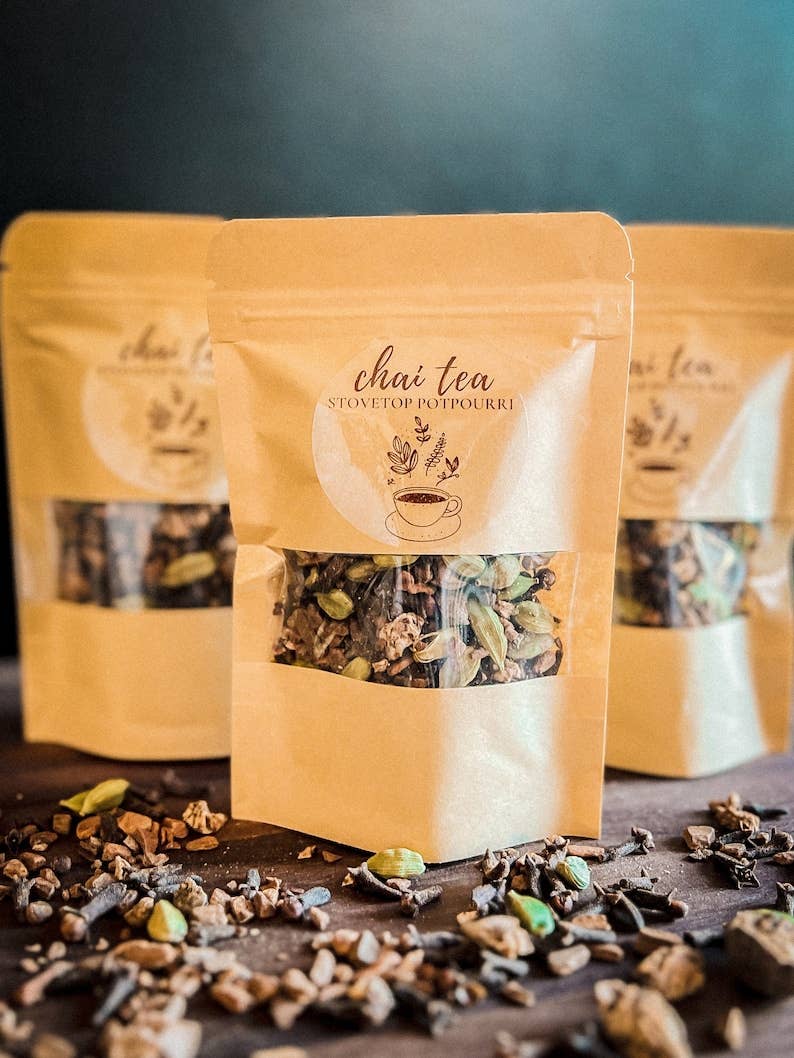 Three stand-up pouches of chai potpourri with a clear window showing loose tea blend, arranged on a dark surface with scattered tea pieces in front. The center pouch is in focus, with the other two blurred in the background.