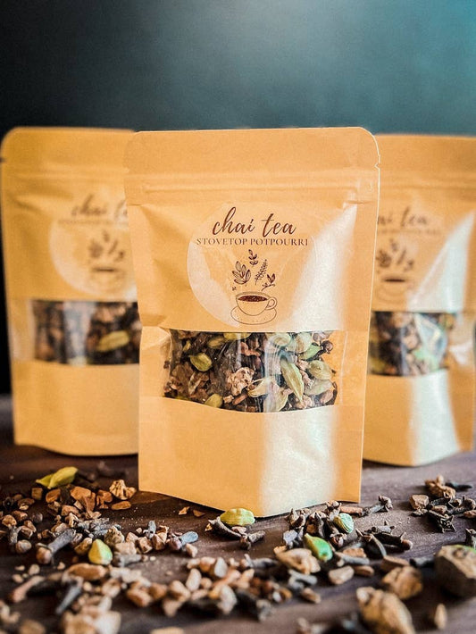 Three stand-up pouches of chai potpourri with a clear window showing loose tea blend, arranged on a dark surface with scattered tea pieces in front. The center pouch is in focus, with the other two blurred in the background.