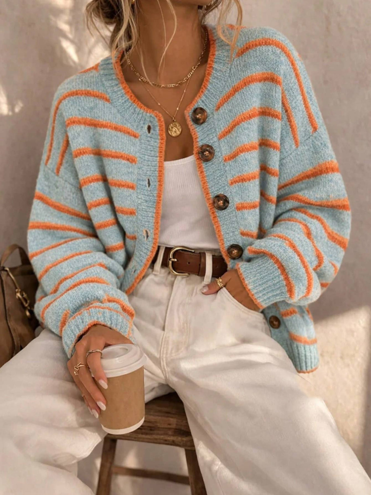 A woman wearing a light blue and orange striped knit cardigan over a white top and white pants, holding a coffee cup while seated on a wooden stool.