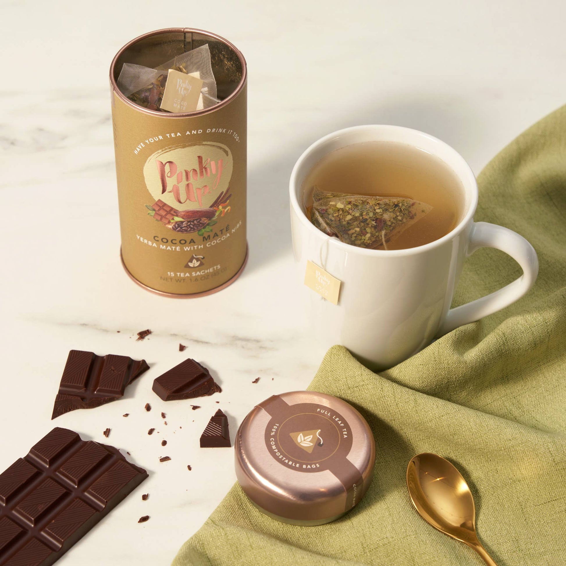 Tea canister, cup of tea with a tea bag, chocolate pieces, and a spoon on a light surface.