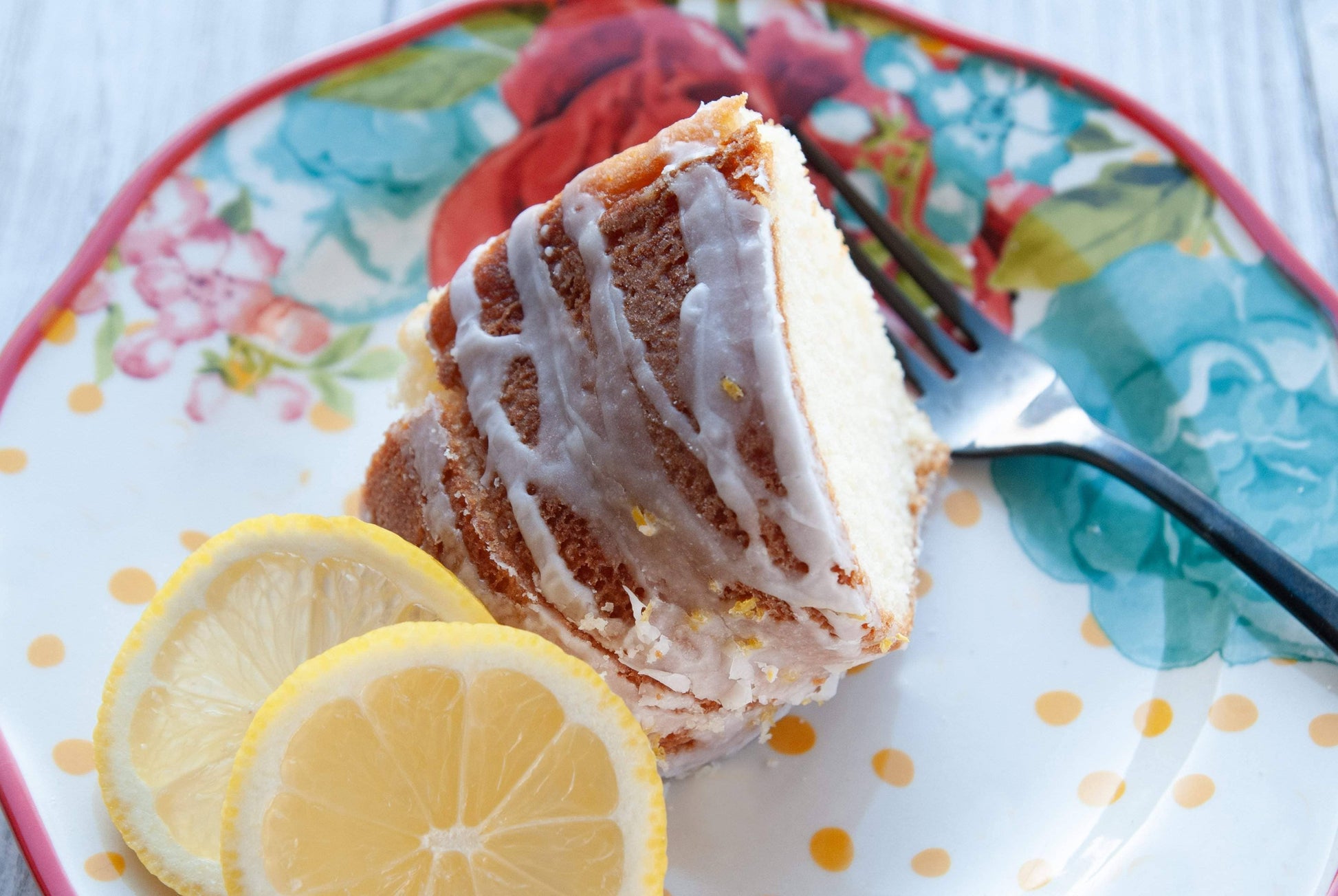 Slice of lemon cake with icing on a decorative plate with lemon slices and a fork.