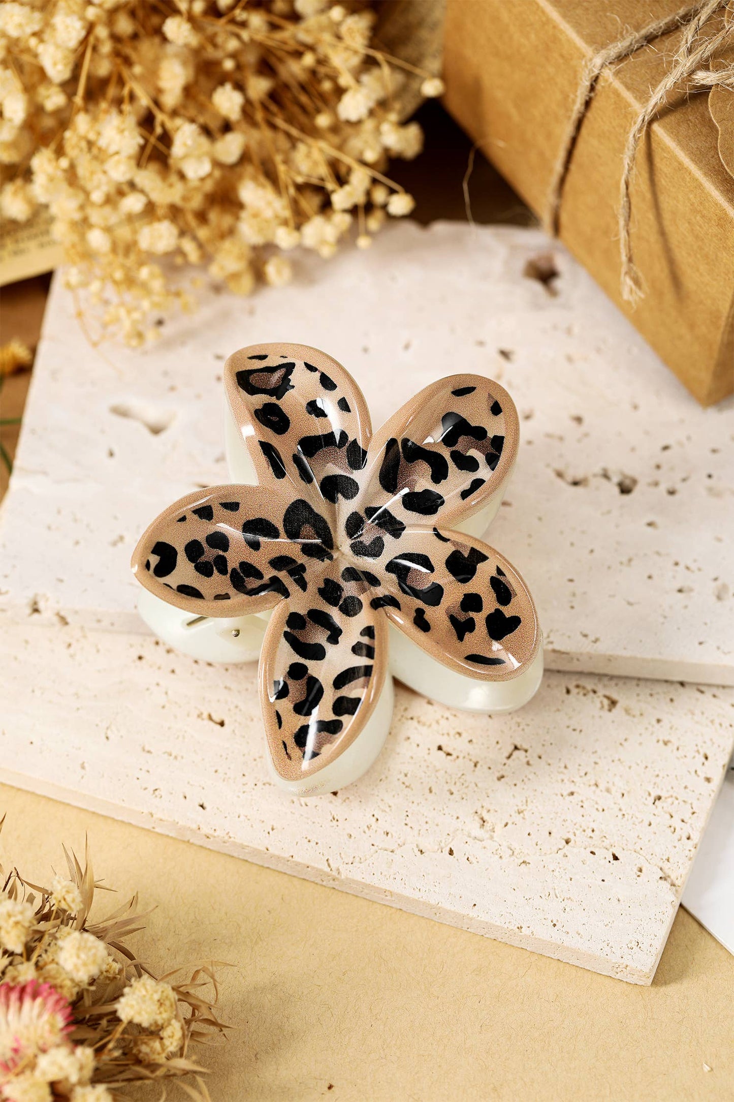 Leopard print flower-shaped hair clip on a textured surface with dried flowers and a brown box in the background.