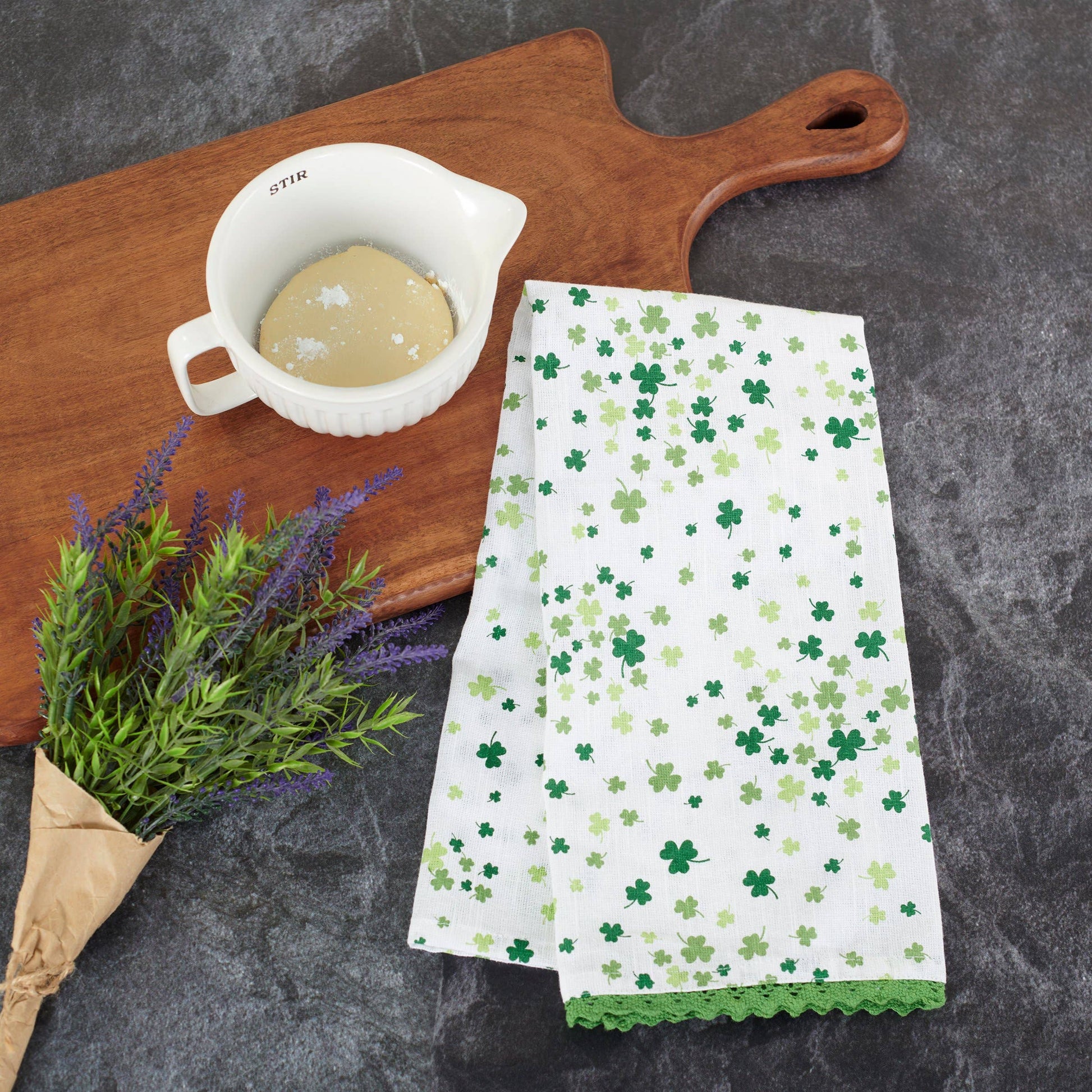 White towel with green shamrock pattern on a dark surface with a wooden cutting board and white cup.