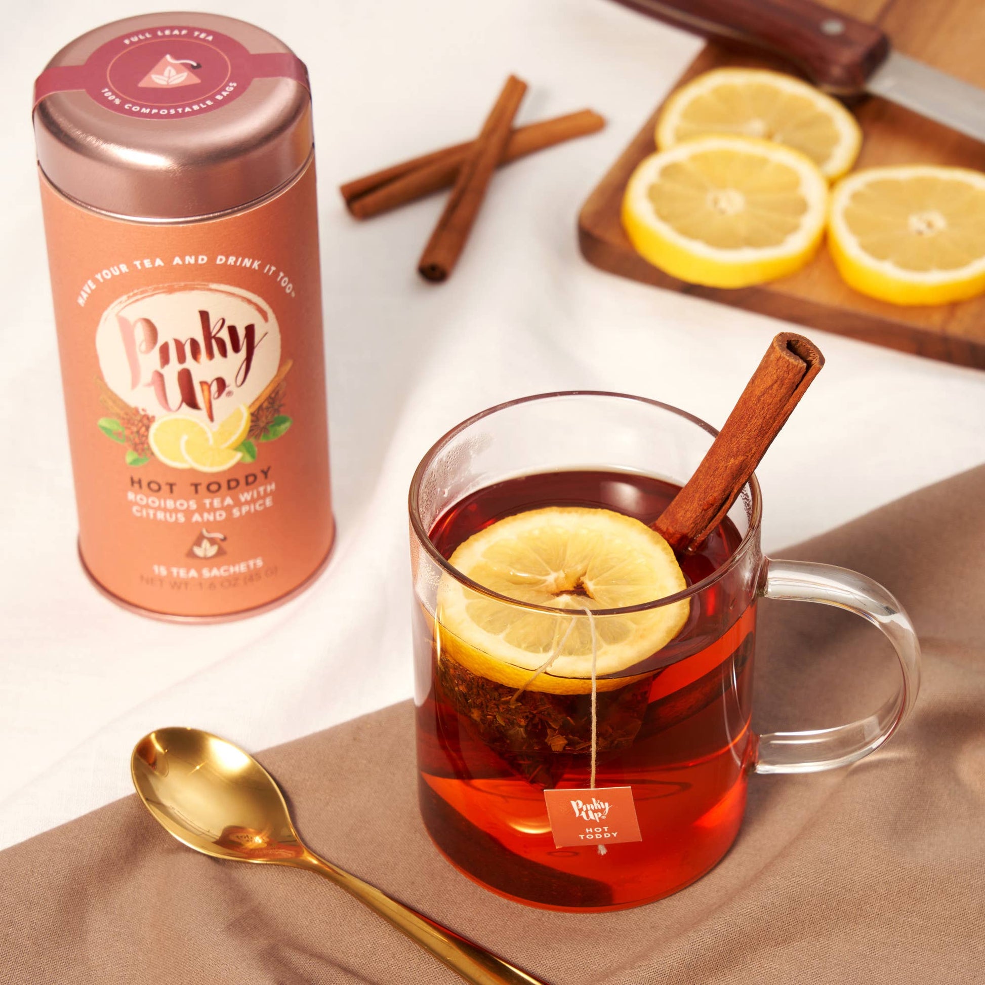 Hot toddy with lemon slice and cinnamon stick, Pinky Up tea packaging, and sliced lemons on a wooden board.