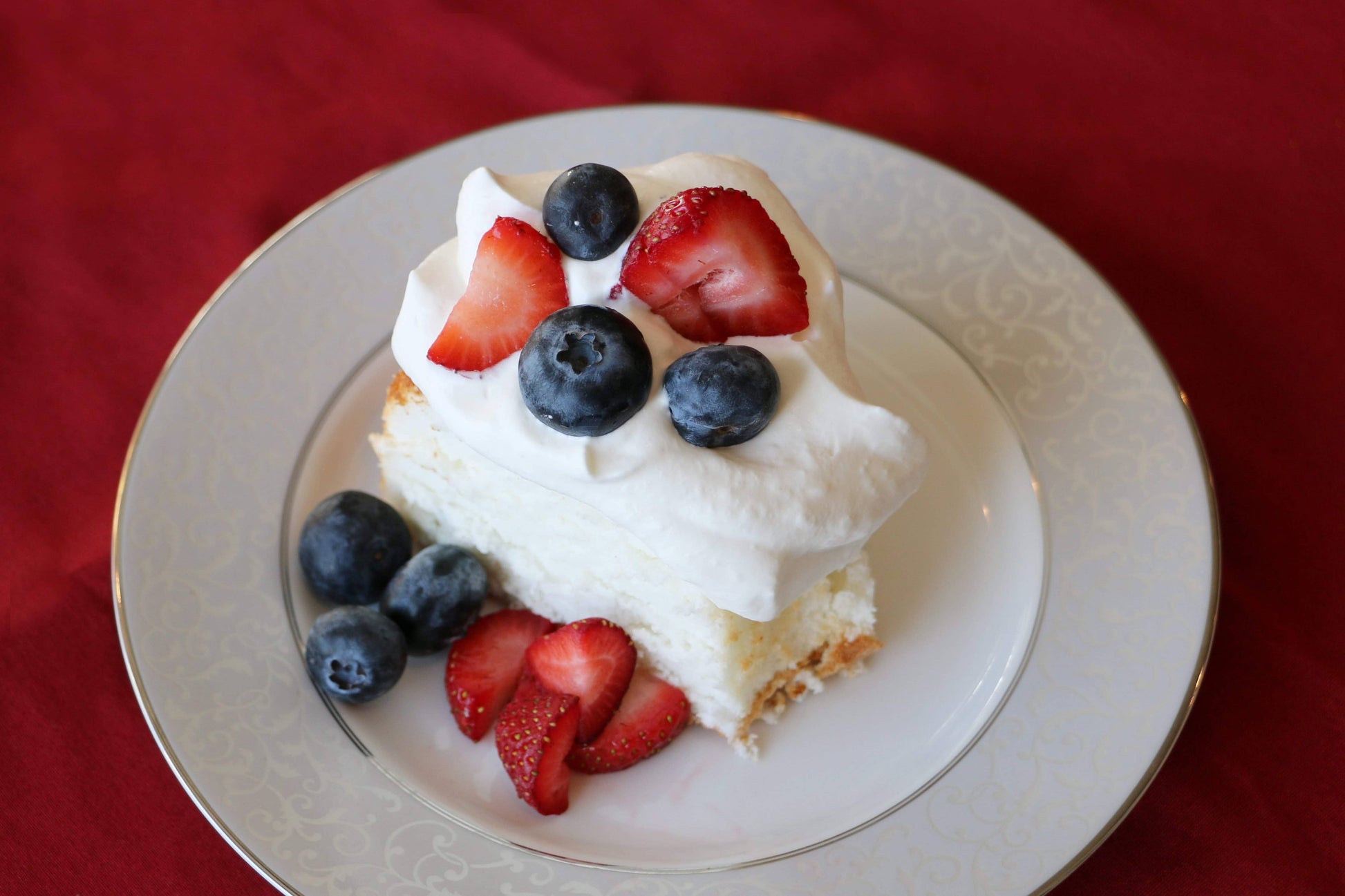 Slice of cake with whipped cream and berries on a decorative plate against a red background