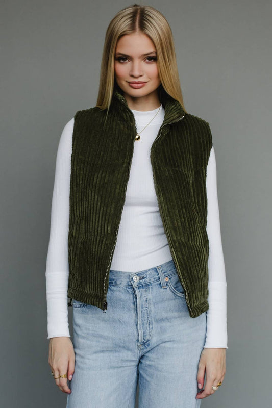 Woman wearing a green corduroy vest over a white shirt and blue jeans on a gray background