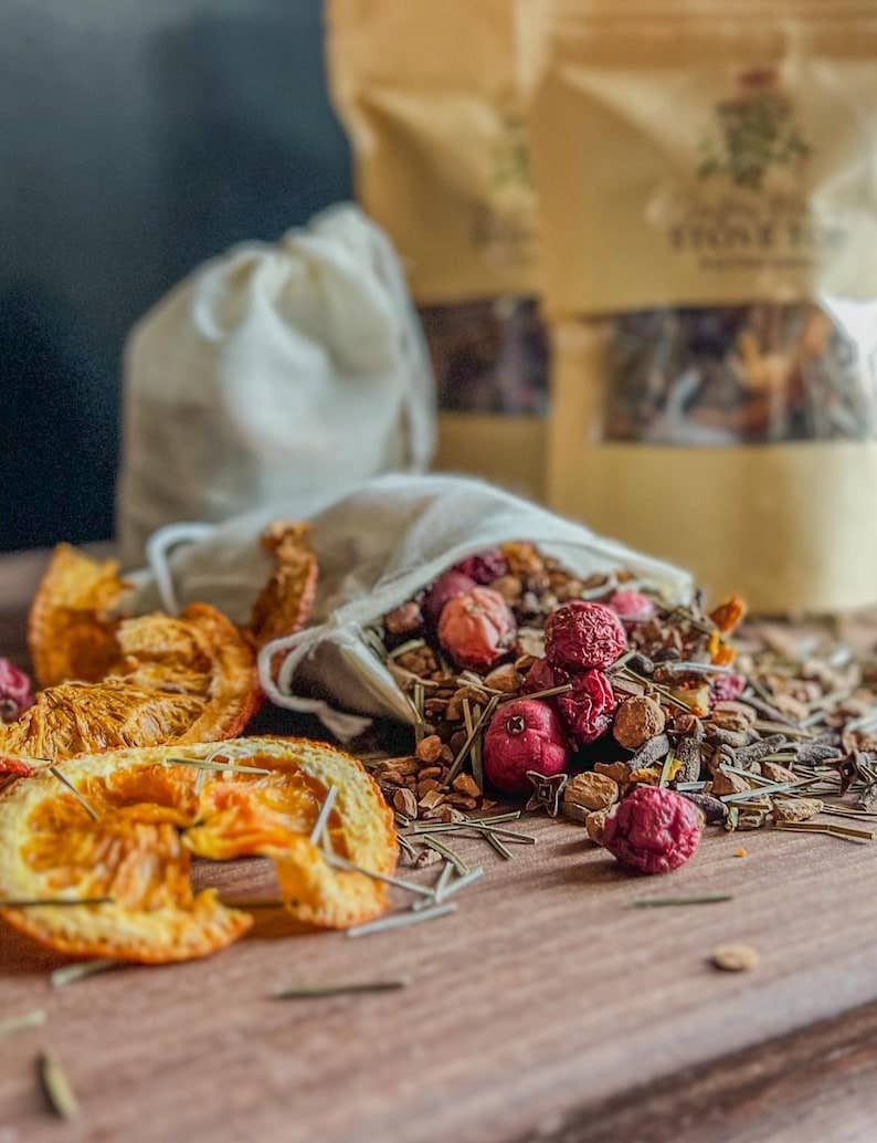 A styled product shot of a dried fruit and herb blend. In the foreground, a small off-white fabric drawstring bag spilling out a colorful mix of dried raspberries, orange slices, and herbs sits on a wooden surface.