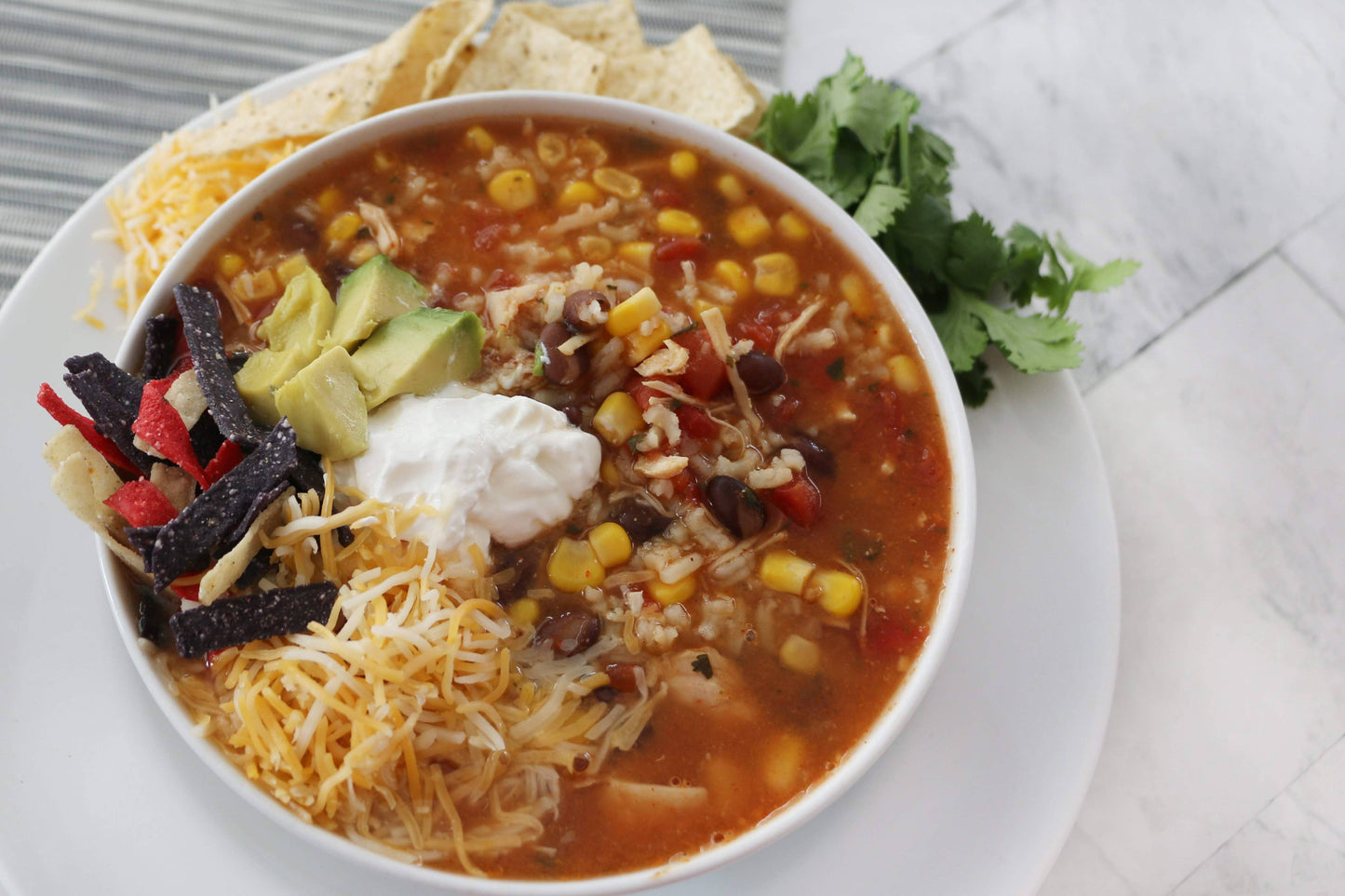 Bowl of taco soup with tortilla strips, cheese, and avocado on a white plate.
