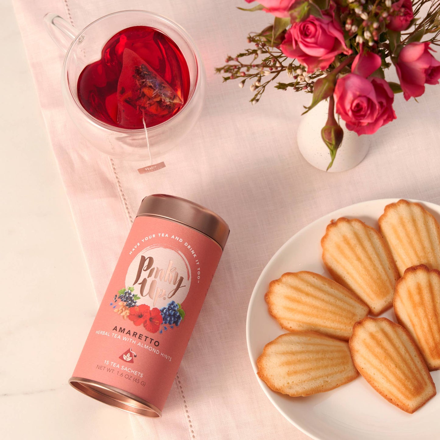 Tea canister with a heart-shaped glass of red tea, cookies, and pink roses on a light surface.