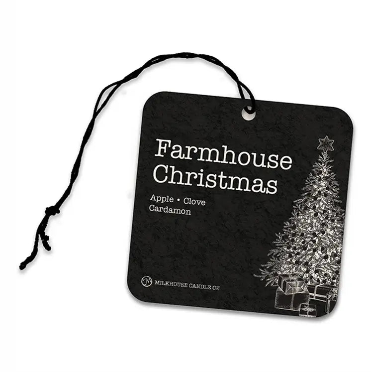 A square black air freshner with rounded corners, a black string loop at the top, and white text reading 'Farmhouse Christmas' plus 'Apple + Clove Cardamon.' A white Christmas tree and gift boxes appear on the right side.
