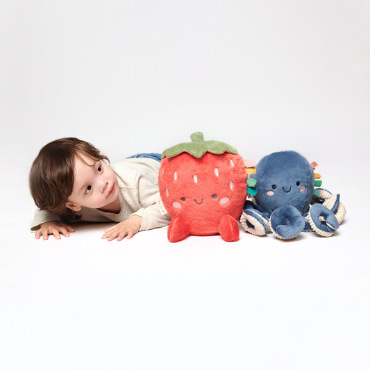 Child lying on a white surface with a strawberry-shaped plush toy and a blue plush toy.