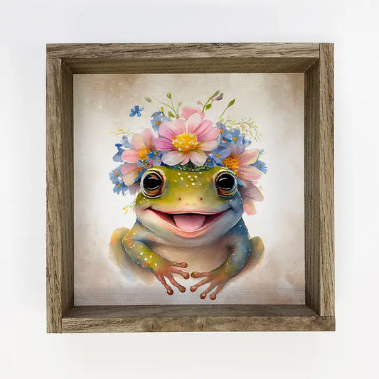 A framed picture of a frog with a floral headband on a white background