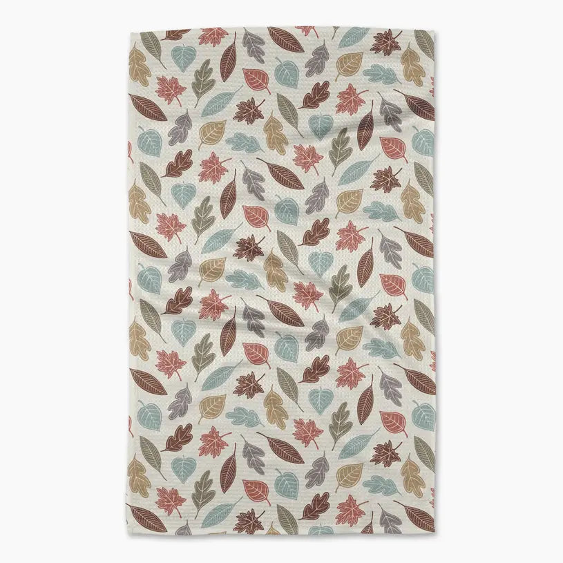 A rectangular kitchen towel with a repeating pattern of autumn leaves in muted shades of red, brown, beige, and light blue on an off-white background, shown flat against a light gray backdrop.