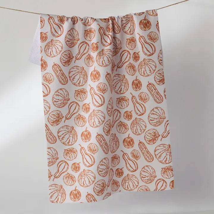 A white kitchen towel or tea towel with an all-over orange pumpkin and gourd print, hanging from a thin string against a plain light gray background.