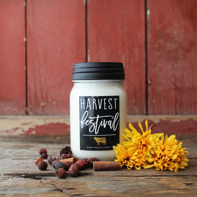 Candle labeled 'Harvest Festival' on a wooden surface with yellow flowers and spices.