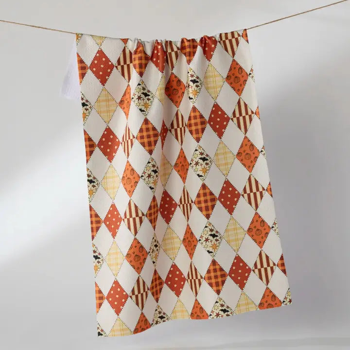 A Geometry Tea Towel with an orange, brown, and cream harlequin diamond pattern, hanging from a thin wire on a light gray background.