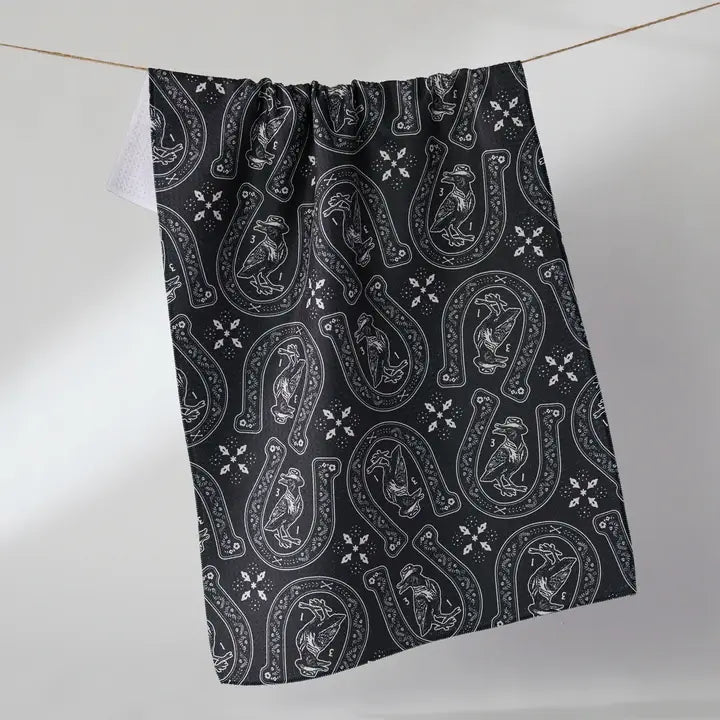 A black kitchen towel with a white horseshoe and chicken pattern, hanging from a string against a light gray background.