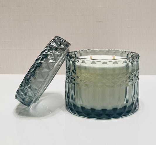 A scented candle with a clear, textured glass jar and a matching glass lid, with two wicks visible on top.
