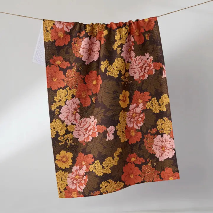 A Geometry Tea Towel with a dense floral pattern in rust orange, pink, and yellow on a dark brown background, hanging from a thin cord against a plain light gray wall.