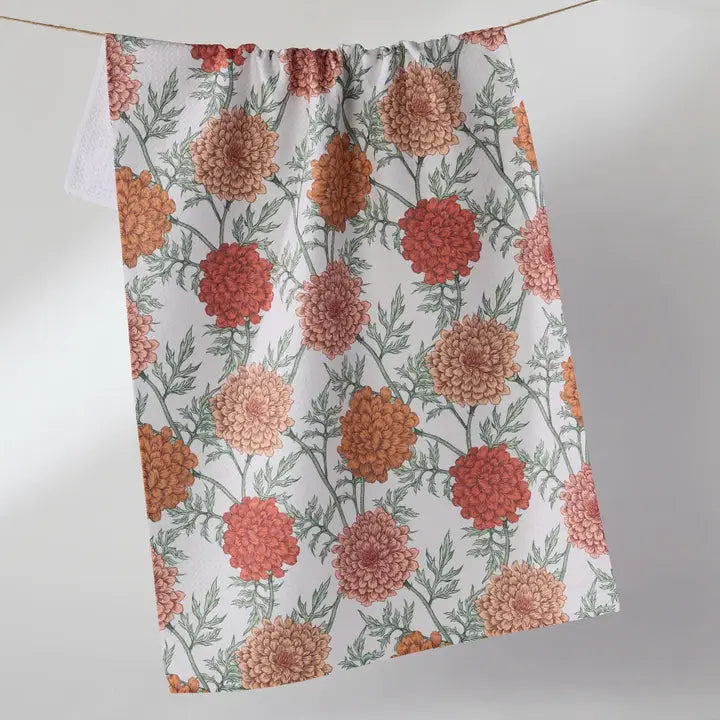 A white Geometry kitchen towel with a repeating floral print of orange, red, and peach marigold flowers with green leaves, hanging from a thin cord against a light gray background.