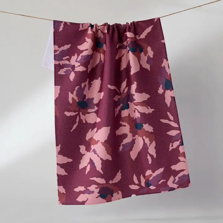 A Geometry tea towel with a floral pattern in dusty pink, mauve, and dark teal, hanging from a thin wire against a plain light gray wall.