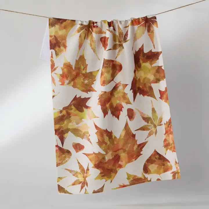 A white kitchen towel featuring a watercolor autumn maple leaf pattern in shades of orange, brown, and yellow, hanging from a string against a light gray background.