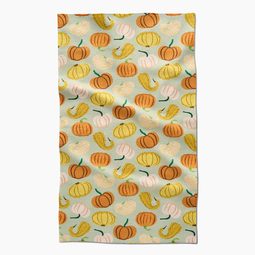 A rectangular kitchen towel with a repeating autumn pattern of orange, yellow, and white pumpkins and gourds on a pale sage green background, shown flat against a light gray backdrop.