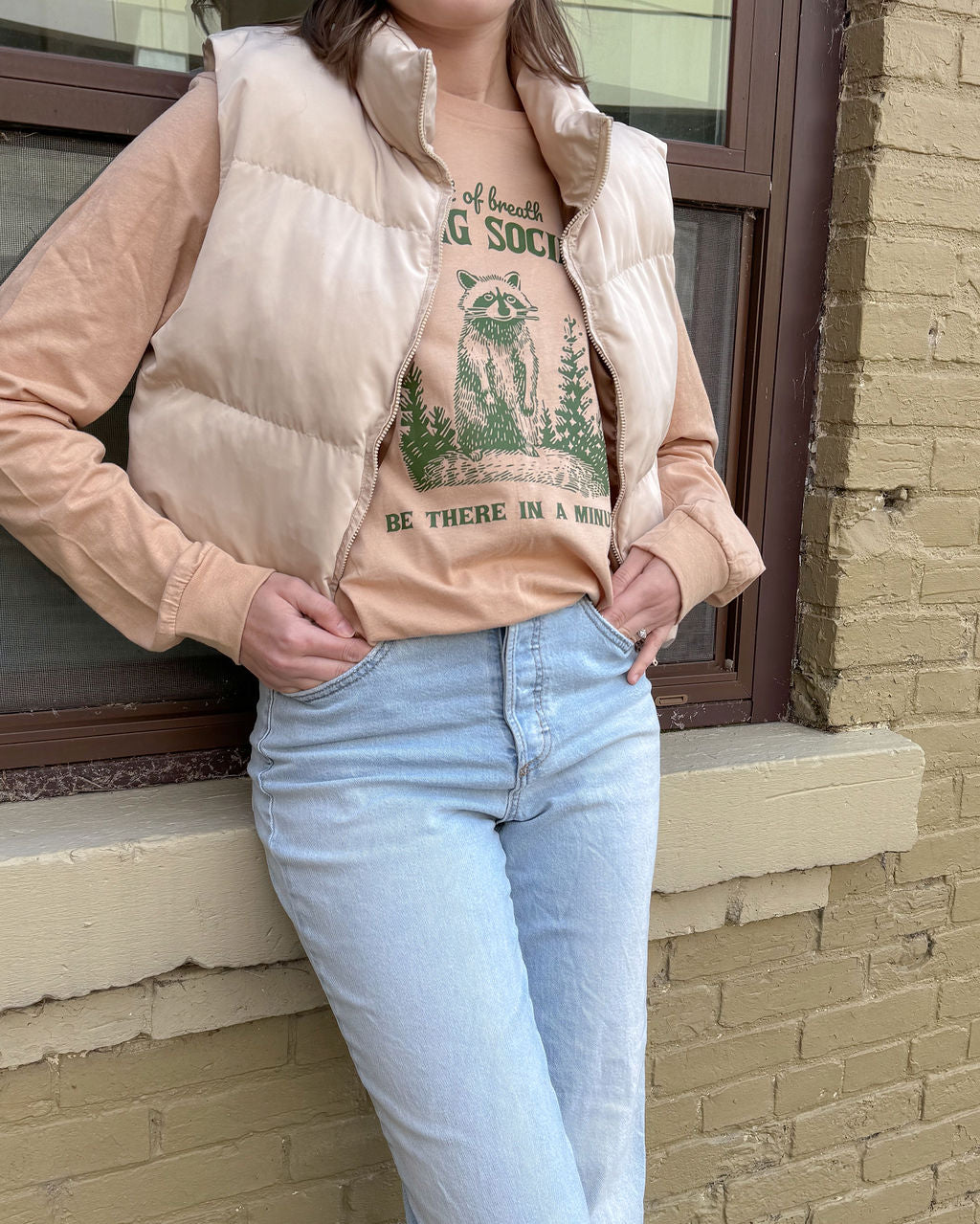 Person wearing a beige puffer vest over a graphic t-shirt with raccoon hiking society funny tee and light blue jeans, standing against a brick wall.