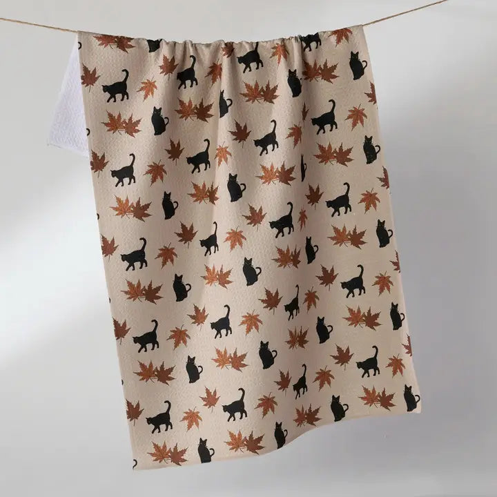 A beige Geometry tea towel  hanging from a thin line, featuring a repeating pattern of black cat silhouettes and brown and orange autumn maple leaves.