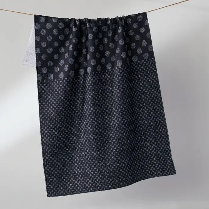 A dark charcoal gray Geometry tea towel with a repeating silver-gray geometric pattern, hanging from a thin rope against a light gray background.