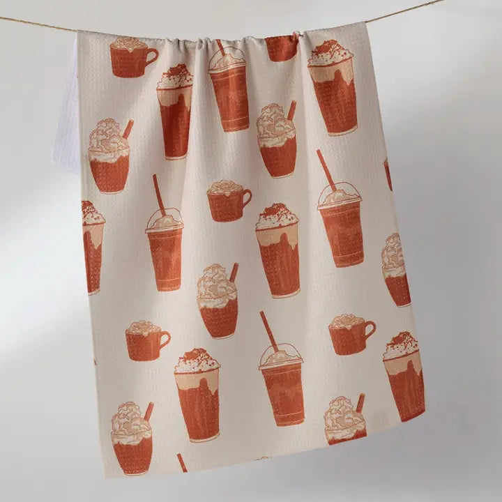 A kitchen towel with a repeating pattern of coffee and milk drinks in terracotta orange and brown on an off-white background, hanging from a string against a neutral gray background.