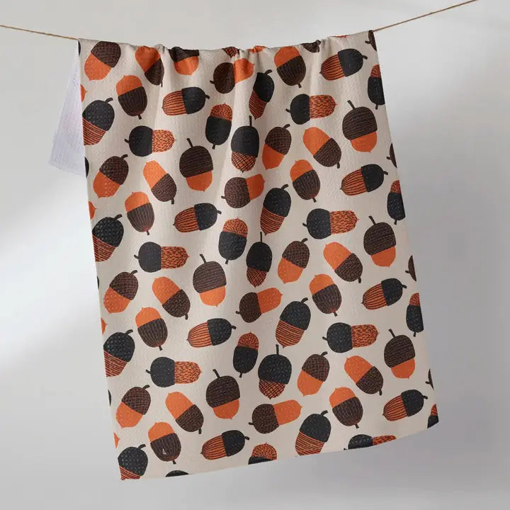A kitchen tea towel with an acorn pattern, hanging from a string against a neutral gray background.